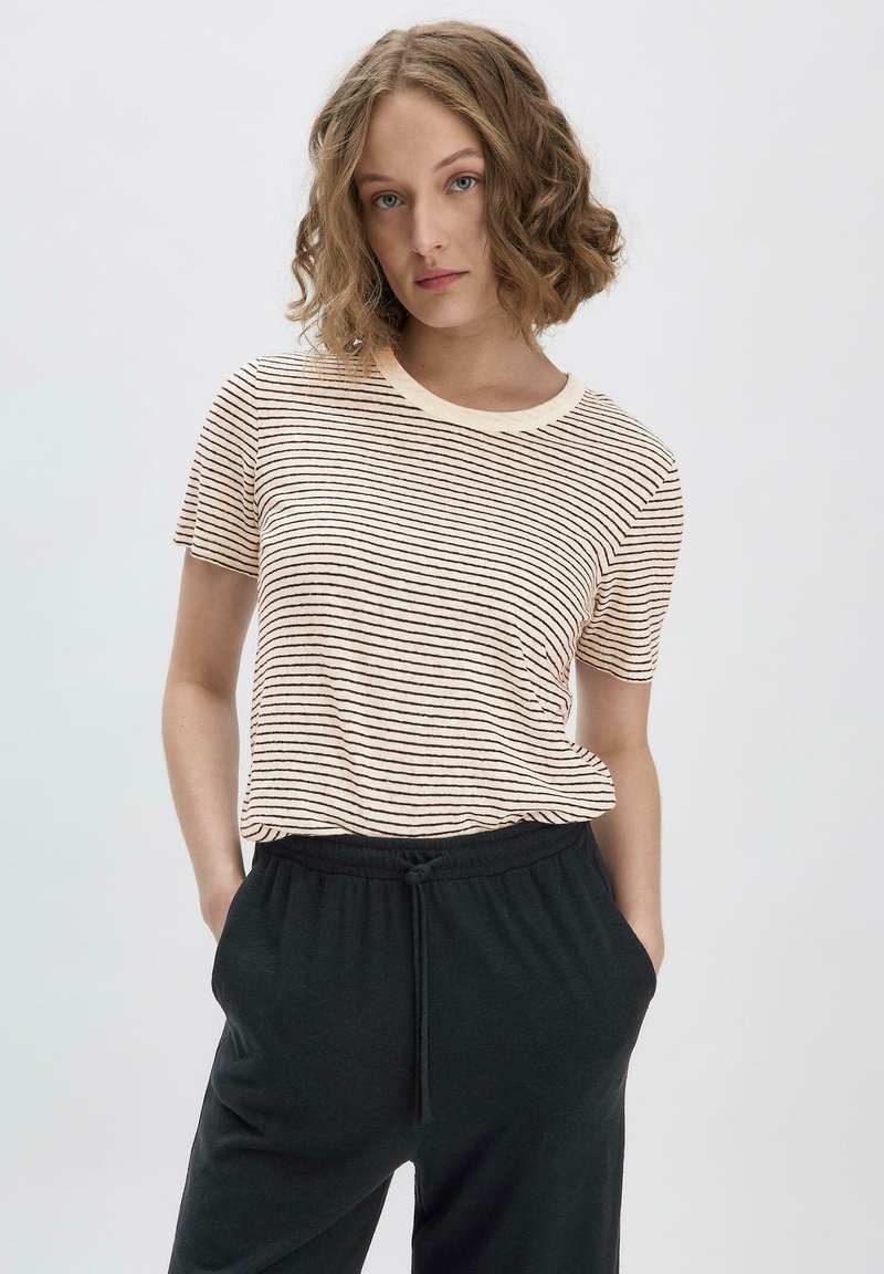 Kerman t-paita, jossa on vaalean kermanväriset ja ruskeat vaakaraitat, pyöreä kaula-aukko, yhdistetty mustien naruvarustesateenkaarihousujen kanssa.