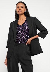 Blazer negro con mangas tres cuartos, combinado con una blusa morada de lentejuelas, con escote en V y detalles que reflejan la luz. Pantalones sastre.