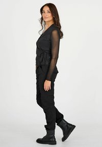 Woman standing sideways wearing sheer long-sleeve black top, black cargo pants, and black lace-up boots against plain white background.