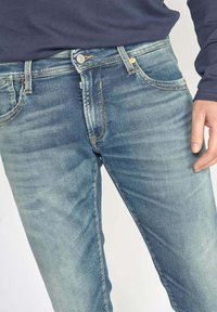 Jeans en denim bleu délavé avec une fermeture éclair, design à cinq poches et coutures contrastées. Vue en gros plan, montrant la texture et l'ajustement.
