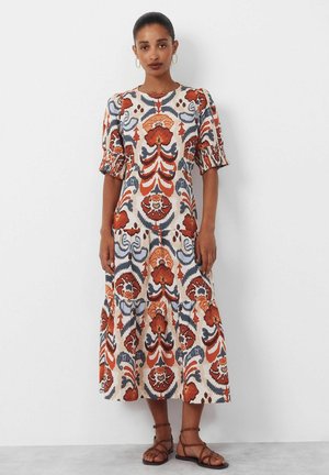 Frau trägt ein Midikleid mit Puffärmeln und einem abstrakten Blumenmuster in Orange, Blau und Beige, kombiniert mit braunen Riemchensandalen.
