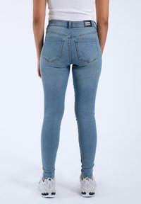 Jeans de mezclilla azul ajustados con cintura alta, que cuenta con dos bolsillos traseros y una etiqueta de la marca. Llevado con zapatillas deportivas blancas y grises.