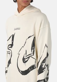 Felpa oversize color crema con audaci stampe grafiche nere sul davanti e sulle maniche, dotata di cappuccio con coulisse e texture morbida.