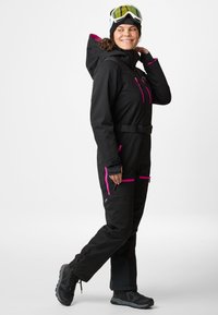Black winter overall with pink accents, features a hood, adjustable belt, and zippered pockets; paired with black boots and ski goggles.