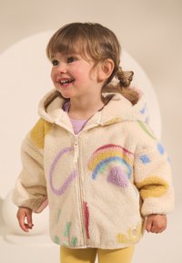 Veste à capuche en polaire couleur crème avec des motifs colorés incluant un arc-en-ciel, des formes géométriques et des accents jaunes. Dotée d'une fermeture éclair frontale.