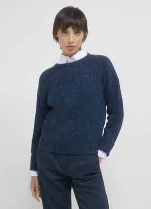 Frau mit kurzem dunklem Haar, die einen navyblauen, gepunkteten Strickpullover über einem weißen Hemd mit Kragen und navyblauen Hosen trägt, vor einem einfarbigen Hintergrund.