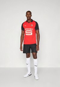 Raudona ir juoda trumpomis rankovėmis futbolo marškinėliai su baltais akcentais, ant kurių didelis skaičius „5“ ir užrašas „SAMSIC“. Juodos kelnės ir baltos kojinės papildo aprangą.