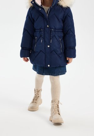 Chaqueta de plumas azul marino con capucha de piel sintética, patrón de rombos, acentos plateados, combinada con una falda de mezclilla, medias crema y botas beige.