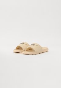 Sandales à glissé beige avec une bande supérieure douce et une semelle texturée, dotées d'une semelle flexible. Branding décoratif noir sur la sangle.