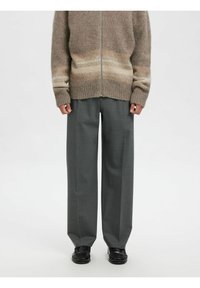 Bēšs un brūns gradienta adīts džemperis ar rāvējslēdzēju un ribotiem manžetēm, kombinēts ar pelēkām plata kleita biksēm un melniem apaviem.