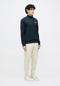 Námornícka bunda na zips s vysokým golierom, s malým logom na hrudi. Kombinovaná s ľahkými béžovými teplákmi a bielymi teniskami.