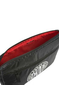 Bolsa de nailon negra con un logo blanco, cierre de cremallera y un interior rojo vibrante con un patrón sutil. Textura suave en toda la superficie.