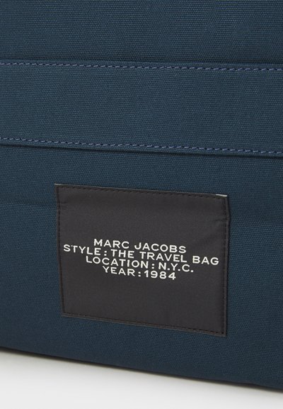 Saco de viagem em lona azul-marinho com costura horizontal, um rótulo preto com texto branco que indica a marca, o estilo, a localização e o ano.