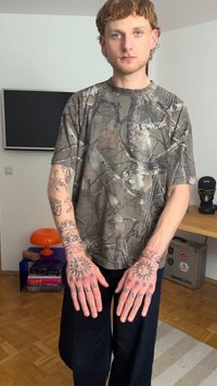 Kameele mustritega lühikeste varrukatega särk tuhmrohelises ja pruunis toonis, kombineeritud mustade pükstega. Kätele on tätoveeritud detailne päikese ja teksti kujundus.