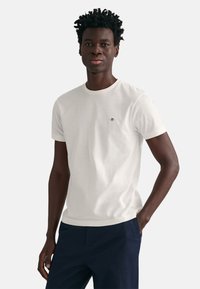 Hvid bomulds T-shirt med rund hals, korte ærmer og et lille broderet logo på brystet, parret med mørke bukser.
