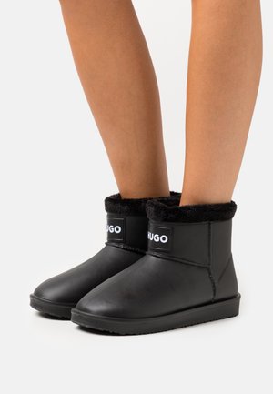 Gambe che indossano stivali neri alla caviglia con fodera pelosa e etichetta "HUGO" sul davanti, su uno sfondo bianco uniforme.