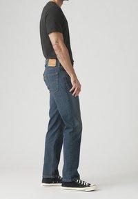 Homme portant un jean bleu foncé, un t-shirt noir et des baskets basses noires, debout de profil sur un fond clair uni.