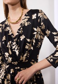 Femme portant une robe noire avec un motif floral doré, encolure en V, manches trois-quarts, collier doré épais et bague dorée à la main droite.