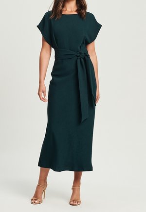 Kobieta ubrana w ciemnozieloną sukienkę midi z krótkimi rękawami i szerokim paskiem związanym na supeł w talii, w połączeniu z beżowymi sandałami na obcasie.