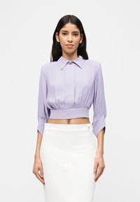 Γυναίκα με μακριά σκούρα μαλλιά, που φοράει ένα λιλά κομμένο μπλουζάκι με φαρδιά γιακά και λευκή φούστα με ψηλή μέση, μπροστά σε απλό φόντο.