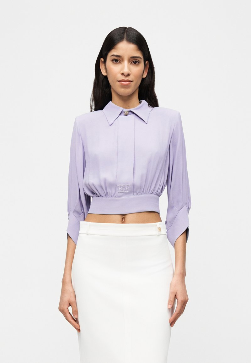 Γυναίκα με μακριά σκούρα μαλλιά, που φοράει ένα λιλά κομμένο μπλουζάκι με φαρδιά γιακά και λευκή φούστα με ψηλή μέση, μπροστά σε απλό φόντο.