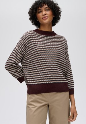 Vrouw met krullend haar, gekleed in een gestreepte bruine, witte en beige gebreide trui en beige broek, staand met één hand op haar heup.