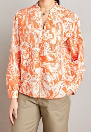 Femme portant un chemisier ample à motif floral orange et blanc avec des manches longues et un pantalon beige, debout avec une main dans la poche.