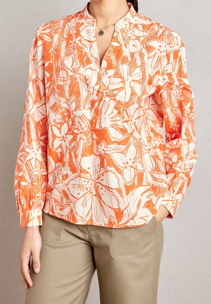 Femme portant un chemisier ample à motif floral orange et blanc avec des manches longues et un pantalon beige, debout avec une main dans la poche.