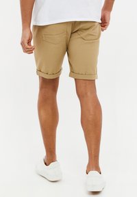 Pantalones cortos color beige con puños enrollados, que cuentan con dos bolsillos traseros. Se combinan con una camiseta blanca y zapatillas blancas sin cordones, sobre un fondo claro.