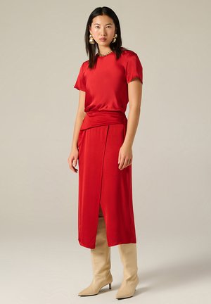 Top in raso rosso con maniche corte abbinato a una gonna avvolgente rossa con spacco laterale, completato da stivali beige con punta a punta al ginocchio.