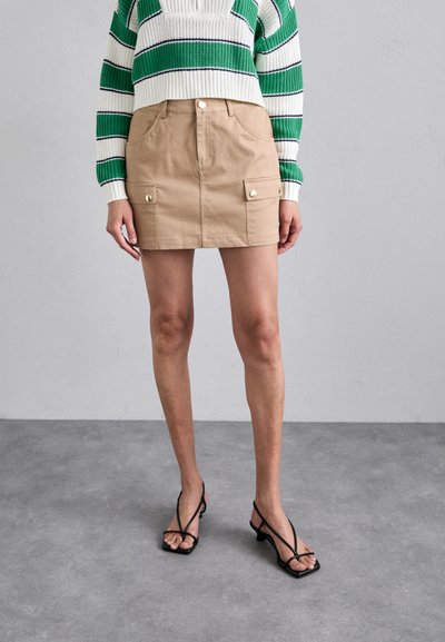 Beige puuvillane mini-seelik esipocketide ja nööbiga, koos rohelise ja valge triibulise kampsuniga. Mustad rihmadega sandaalid.