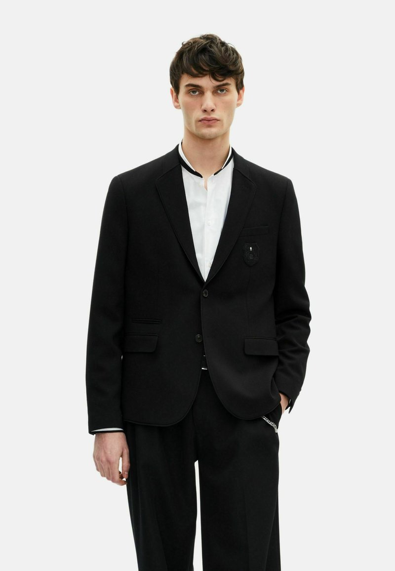 Schwarzer Wollblazer mit strukturiertem Design, Reverskragen und einer vorderen Tasche mit einer Paspel. Kombiniert mit einem weißen Hemd.