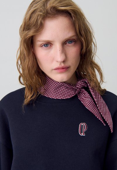 Sudadera navy con el logo bordado 'CP', combinada con una bufanda de cuadros burdeos atada al cuello. Textura suave y diseño informal.