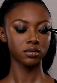 Close-up of a person applying makeup; features dark smoky eyeshadow, glossy lips, smooth skin, and dark hair. Cosmetic brush in use.