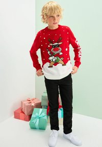 Rød julesweater med en tegneserie-rensdyr i solbriller, snefnug og en hvid nederste sektion; parret med sorte bukser og hvide sneakers.