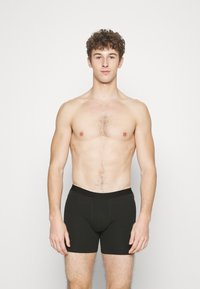 Black cotton boxer shorts with a snug waistband, featuring a smooth texture and a front pouch for support. The model stands upright.