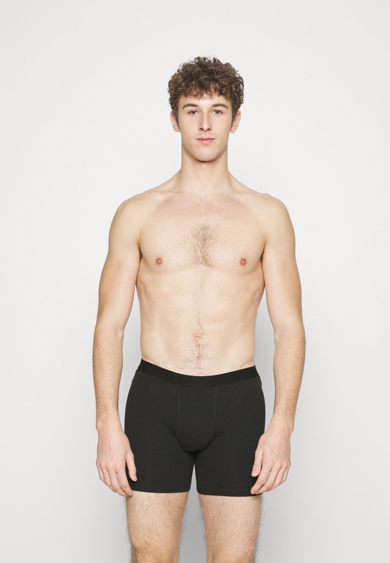 Black cotton boxer shorts with a snug waistband, featuring a smooth texture and a front pouch for support. The model stands upright.
