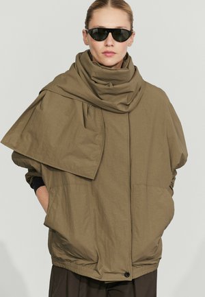 Kvinde iført oversized khakifarvet jakke med høj krave og draperet stofdetalje, sorte solbriller, hænder i lommerne, neutralt baggrund.