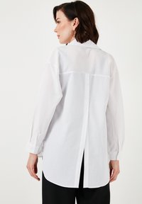 White long-sleeve shirt with a back yoke, button-down closure, and a split hem. Made from lightweight fabric with a smooth texture.