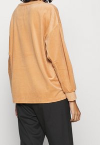 Persoon met een oversized, ribbelige, lichtbruine corduroy lange mouw shirt en zwarte broek, weergegeven van de achterkant en zijkant.