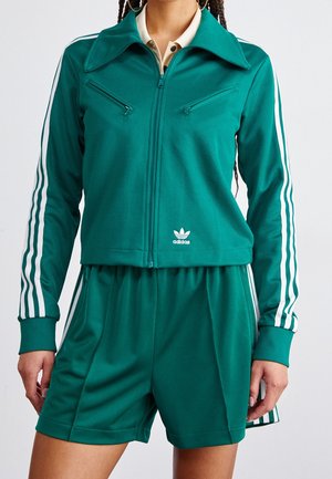 Kobieta w zielonej kurtce sportowej Adidas z białymi paskami na rękawach oraz dopasowanych szortach z białymi paskami po bokach.