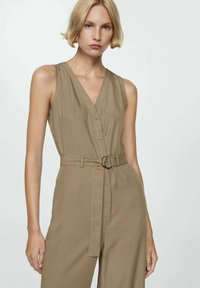 Beige ärmlös jumpsuit med djup V-ringning, knapprad framtill och belagd midja med en metallring. Tillverkad av mjukt tyg med en slät textur.