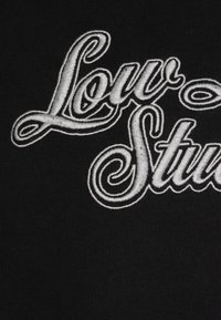 Tissu noir avec texte brodé blanc indiquant "Low Studio" dans une police décorative courbée avec des détails complexes.