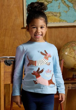 Usmívající se dítě s kudrnatými vlasy má na sobě světle modrou košili s motivem dvou jezevčíků v svetrech a s brýlemi ve tvaru srdce, stojící ve třídě.