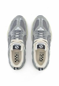 Zapatillas de malla y gamuza grises con acentos en blanco roto. Suela gruesa con logo negro y patrón de tablero de ajedrez en la lengüeta. Textura de la suela de goma visible.