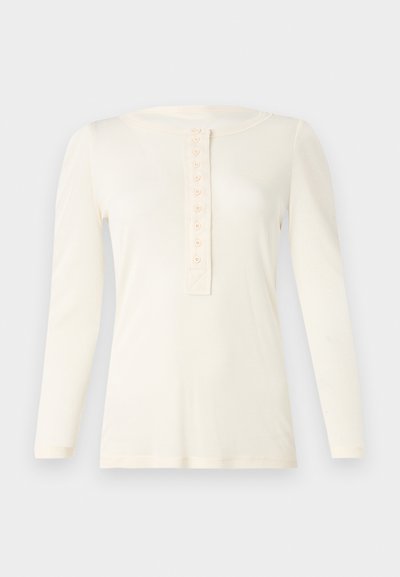 Béžová blúzka s dlhým rukávom vyrobená z mäkkej tkaniny, ktorá má okrúhly výstrih a légu s deviatimi gombíkmi uprostred.