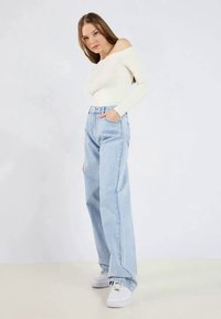 Bela off-shoulder majica v kombinaciji s svetlo modrimi visokimi širokimi hlačami. Obleka dopolnjena z belimi supergami, z rokama v žepih.