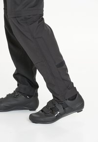 Mustat pyöräilykengät, joissa on kolme tarranauhaa ja rei'itettyjä yksityiskohtia, yhdistettynä tummiin, teksturoiduihin, istuviin housuihin, joissa on tarranauha yksityiskohtia.