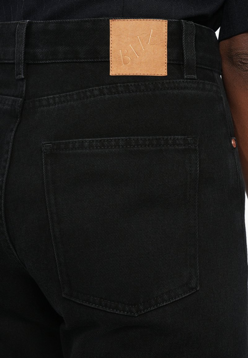 Jean en denim noir avec une étiquette en cuir sur la taille. Dispose de poches arrière classiques avec des détails de couture subtils. Texture de tissu durable.