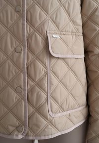 Quiltet beige jakke med diamantmønster, kontrastkant, trykknapper og en liten frontlomme med merkeetikett.